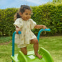 A young girl stands atop the TP Toys 6ft (1.8m) Wavy Slide & Stepset, holding blue handles, with grass and hedges in the background—perfect for any garden playground.