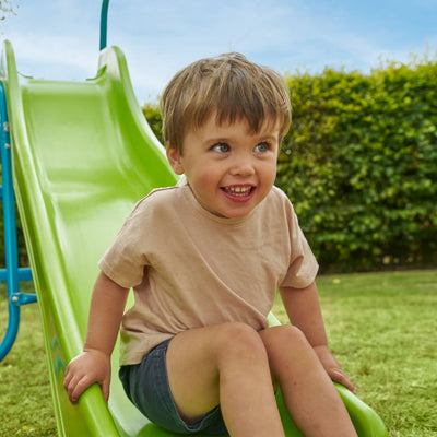 A young child smiles while sitting at the top of the TP Toys TP 6ft (1.8m) Wavy Slide & Stepset in a garden playground, looking slightly to the side.