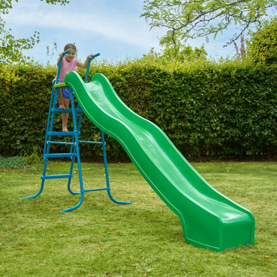 A child stands atop the TP Toys 10ft (3m) Super Wavy Slide & Stepset with blue steps in a grassy backyard, ready for an adventure among tall hedges.