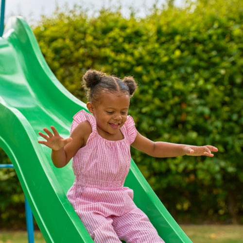 A young girl in a pink-striped outfit slides down the TP Toys 10ft (3m) Super Wavy Slide & Stepset at an adventure playground, with green bushes in the background.