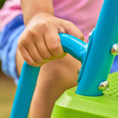 A child grips a blue metal bar on TP Toys' green structure, their colorful clothes visible, just before zooming down the TP 10ft (3m) Super Wavy Slide & Stepset.