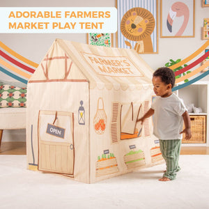 A young child stands beside the TP Toys Activo Farmers Market Play Tent, an imaginative indoor-outdoor playhouse with colorful produce graphics and an open sign.