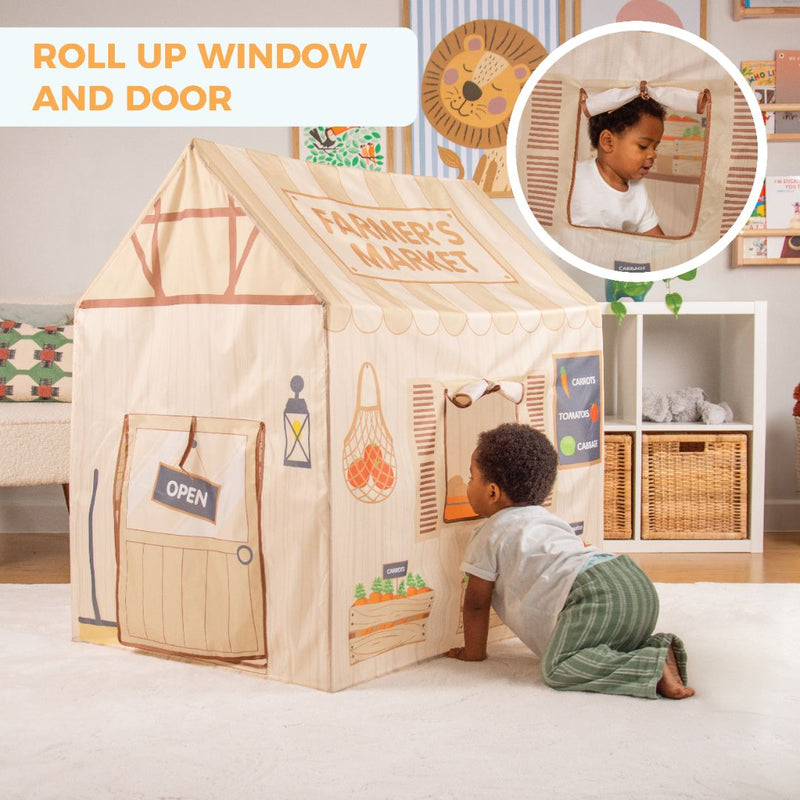 A child peers into the TP Toys Activo Farmers Market Play Tent, a playhouse with a roll-up window and door. Set up indoors or outdoors in the playroom, it’s surrounded by storage baskets and toys.
