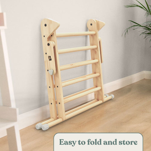 A folded TP Toys TP Active-Tots Pikler Style Wooden Climbing Triangle & Slide (FSC® certified) leans against a wall on a hardwood floor, with a plant in the background. Text at the bottom reads: Easy to fold and store.