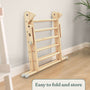 A folded TP Toys TP Active-Tots Pikler Style Wooden Climbing Triangle & Slide (FSC® certified) leans against a wall on a hardwood floor, with a plant in the background. Text at the bottom reads: Easy to fold and store.