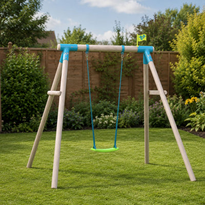 The TP Toys TP Sherwood Single Wooden Swing Set, featuring a PEFC certified roundwood frame and blue metal brackets, stands on a green lawn surrounded by bushes and a wooden fence beneath a partly cloudy sky.