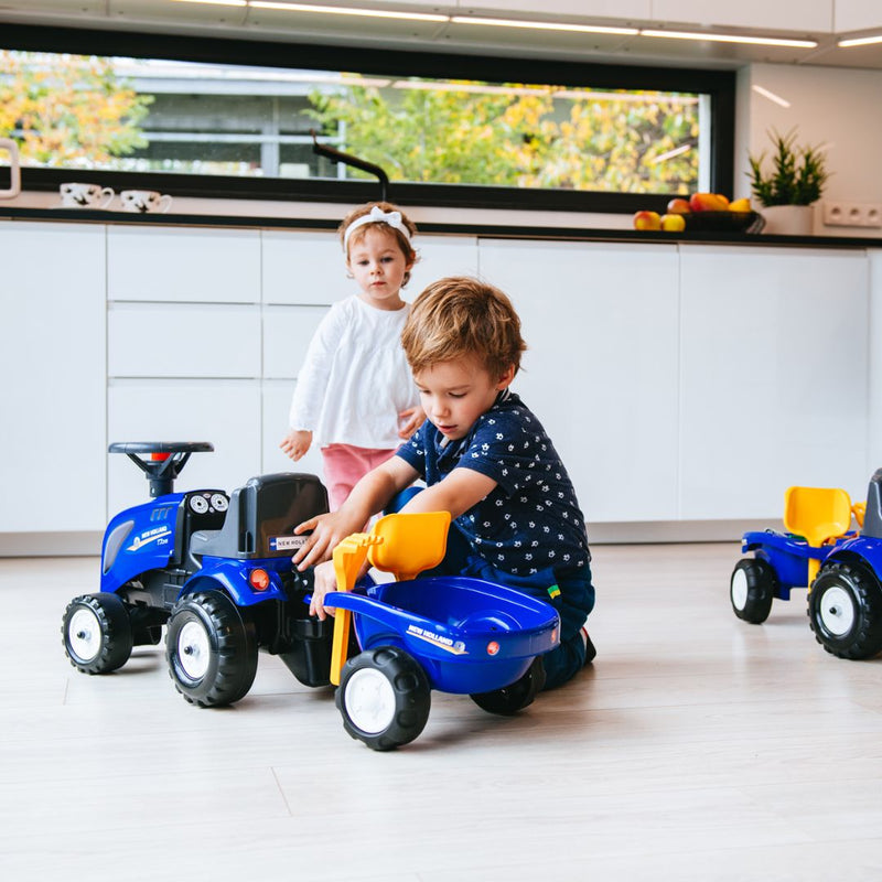 A young boy plays with the TP Toys Falk New Holland Ride-on Tractor with Trailer, Rake & Shovel on the kitchen floor, boosting motor skills, while a young girl stands nearby, watching him.