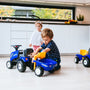 A young boy plays with the TP Toys Falk New Holland Ride-on Tractor with Trailer, Rake & Shovel on the kitchen floor, boosting motor skills, while a young girl stands nearby, watching him.