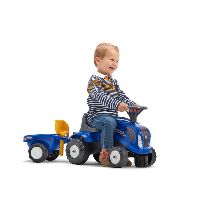 A young child smiles while riding the TP Toys Falk New Holland Ride-on Tractor with Trailer, Rake & Shovel, ideal for developing motor skills, against a white background.