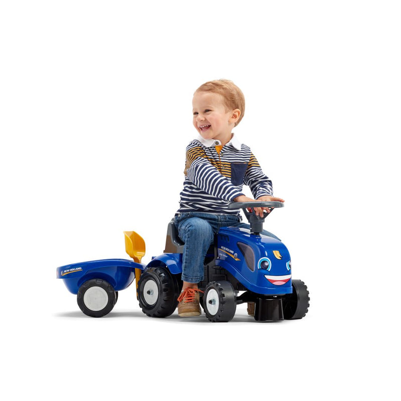 A young child smiles while sitting on the Falk New Holland Ride-on Tractor with Trailer, Rake & Shovel by TP Toys, promoting motor skills development against a white background.