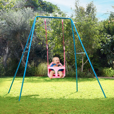 A baby smiles in a sunlit garden, sitting in the pink Quadpod on the TP Toys Small to Tall 2 in 1 Metal Swing Set, surrounded by green grass and trees.