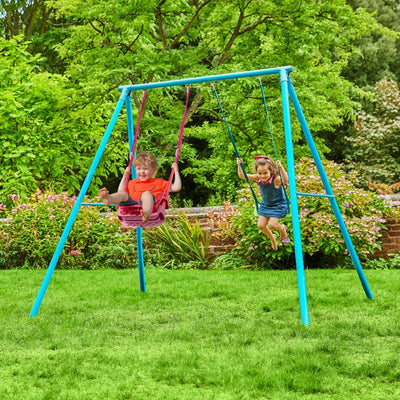 Two young children enjoy the TP Toys Deluxe Double Metal Swing with Rapide and Pink Quadpod seats, featuring a powder-coated steel frame, in a grassy backyard surrounded by green trees and shrubs.