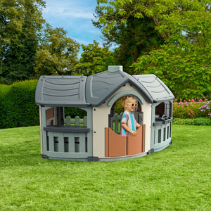 A child enjoys imaginative play on a sunny lawn in the TP Toys Palplay Grand Villa Plastic Playhouse, surrounded by trees and shrubs.
