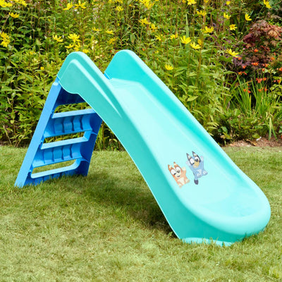The TP Toys Bluey Plastic Folding Slide features cute cartoon dog stickers and sits on grassy ground with yellow flowers—ideal for indoor or outdoor toddler fun.