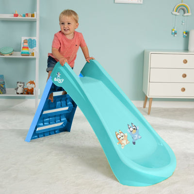 A young child stands atop the TP Toys Bluey Plastic Folding Slide, featuring blue and teal colors with cartoon characters, in a neatly furnished playroom.
