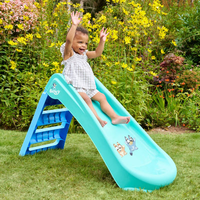 A young child with short hair slides down a TP Toys Bluey Plastic Folding Slide outdoors, smiling with arms raised, surrounded by green grass and yellow flowers.