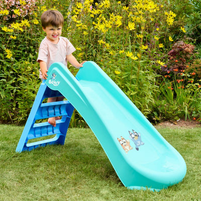 A young child enjoys the TP Toys Bluey Plastic Folding Slide, decorated with Bluey characters and set on garden grass—perfect for indoor or outdoor toddler fun.