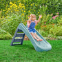 A young child enjoys sliding down the TP Toys PalPlay Misty Sky Plastic 4ft Folding Slide in a garden filled with green grass and colorful flowers, wearing overalls and a striped shirt.