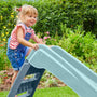 A young girl in denim overalls and a striped shirt climbs up the TP Toys PalPlay Misty Sky Plastic 4ft Folding Slide in a garden filled with green plants and yellow flowers.