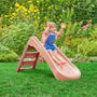 A young child slides down the TP Toys PalPlay Pink Blush Plastic 4ft Folding Slide in a garden with green grass and colorful flowers.