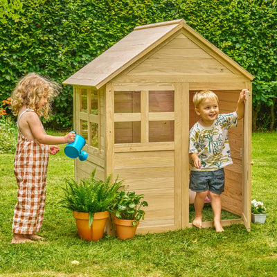 Two young children enjoy a lush yard with the TP Toys Buttercup Cottage Wooden Playhouse (FSC® certified); one waters potted plants while the other stands in the open doorway of the charming structure.