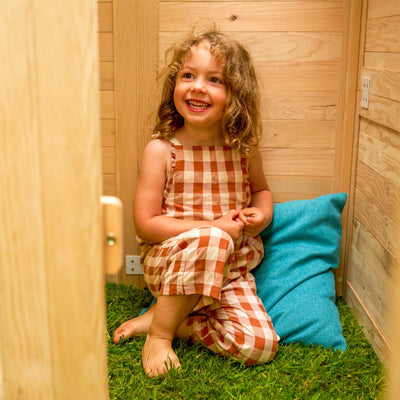 A young child in a gingham outfit smiles beside a blue pillow, sitting on artificial grass inside the Buttercup Cottage Wooden Playhouse - FSC® certified by TP Toys.