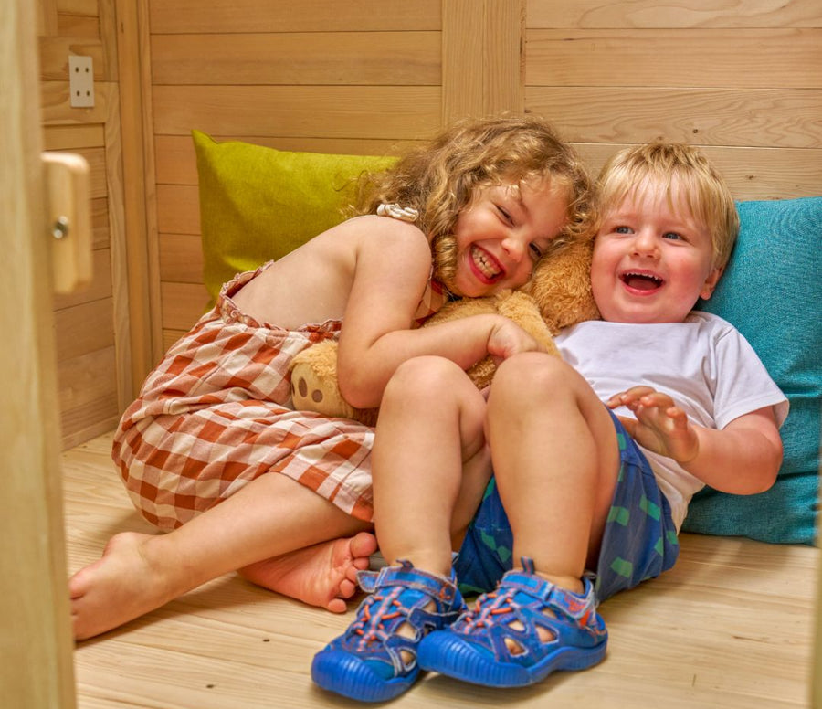 Two young children sit on the floor of the TP Toys Buttercup Cottage Wooden Playhouse with Floor, Verandah and Mud Kitchen, hugging a teddy bear and laughing, colorful pillows behind them.