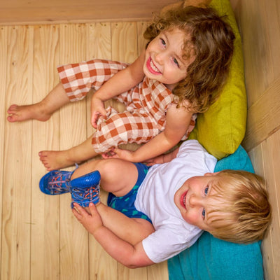 Two young children smile up at the camera as they sit on colourful cushions beside the TP Toys Buttercup Cottage Wooden Playhouse with Floor, Verandah and Mud Kitchen, made from FSC® certified wood.