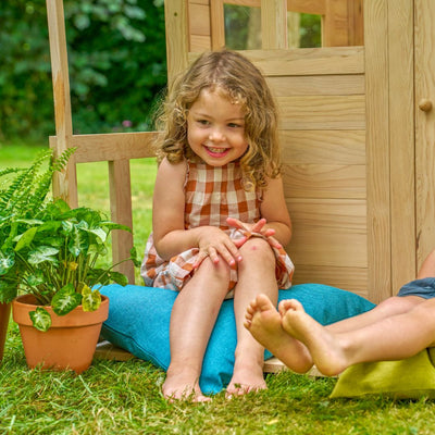 A young child in a checkered dress sits on a blue cushion beside a potted plant and the TP Toys Buttercup Cottage Wooden Playhouse with Floor and Verandah (FSC® certified), smiling as another childs feet appear nearby.