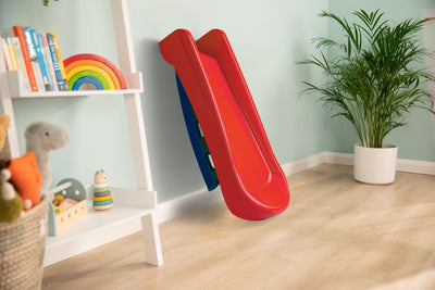 The TP Toys PalPlay Red & Blue Plastic Folding First Slide is set indoors against a light green wall, next to a white toy shelf and potted plant on a wooden floor.