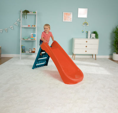 A young child stands atop the TP Toys PalPlay Red & Blue Plastic Folding First Slide in a light blue, neatly decorated room with a white rug and children’s furniture.
