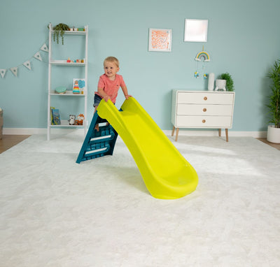 A young child in a red shirt stands at the top of a TP Toys PalPlay Green & Blue Plastic Folding First Slide in a neatly decorated playroom.