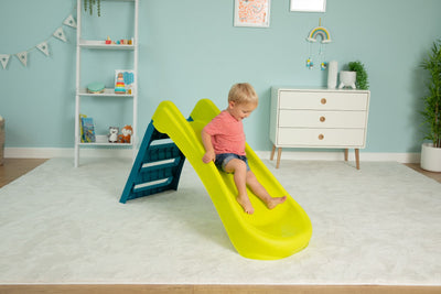 A young child in a red shirt and shorts slides down the TP Toys PalPlay Green & Blue Plastic Folding First Slide in a playroom with light blue walls and white furniture.