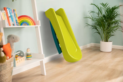 The TP Toys PalPlay Green & Blue Plastic Folding First Slide is set against a light blue wall in a playroom with wooden flooring, a plant, and a bookshelf with toys, adding indoor and outdoor fun for children.