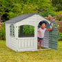 A young child smiles while stepping out of the TP Toys Palplay Plastic Playhouse of Fun Snowdrift, perfect for outdoor use, in a garden with green grass and flowers in the background.