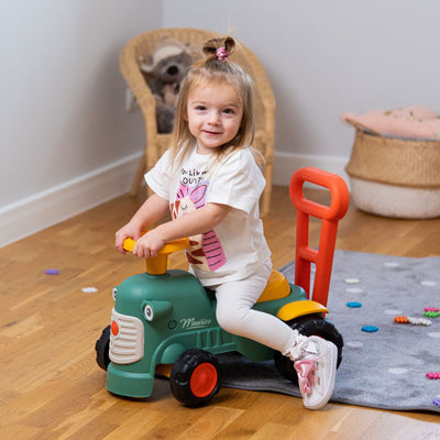 A young girl sits on the Falk Maurice Vintage Baby Green Tractor by TP Toys in a room with a gray rug, wooden floor, and a woven basket in the background.