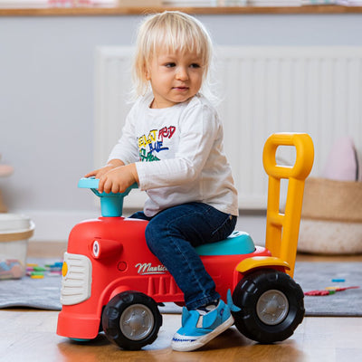A young child with blonde hair sits indoors on a TP Toys Falk Maurice Vintage Baby Red Tractor, wearing a white shirt, blue jeans, and blue shoes.