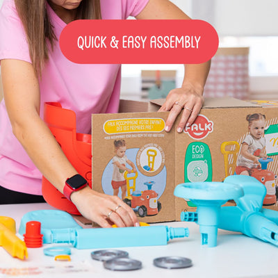 An adult assembles a TP Toys Falk Maurice Vintage Baby Red Tractor from recycled plastic parts, with the walking aid bar, product box, and assembly instructions on the table to help little ones learn to walk.
