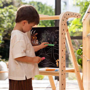 A young boy stands on the TP Toys The Gruffalo Wooden Helper Tower – FSC® certified, drawing a scene with trees and animal characters on a chalkboard easel using colored chalk in a sunlit room.