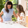 An adult and a child stand at a white table sorting stacks of cards, with sunlight streaming through a window and greenery visible outside. The child stands confidently on the TP Toys Gruffalo Wooden Helper Tower – FSC® certified, which brings them to just the right height for tabletop activities.