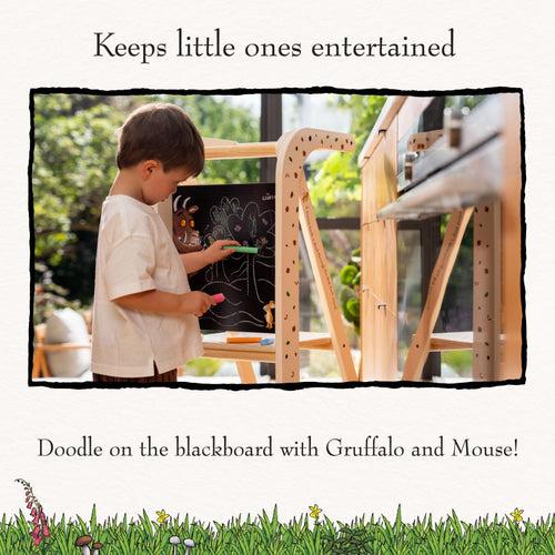 A young child stands confidently on the TP Toys The Gruffalo Wooden Helper Tower - FSC® certified, drawing with colored chalk on a blackboard adorned with charming Gruffalo and Mouse illustrations in a brightly lit room.
