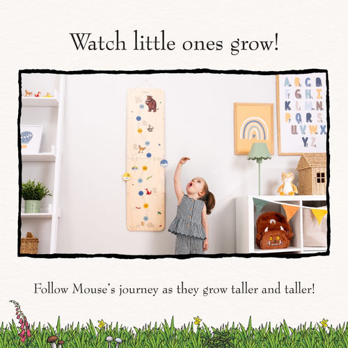 A young child measures their height with The Gruffalo Wooden Height Chart - FSC® certified by TP Toys in a playroom filled with books, toys, and colorful art.