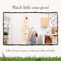 A young child measures their height with The Gruffalo Wooden Height Chart - FSC® certified by TP Toys in a playroom filled with books, toys, and colorful art.