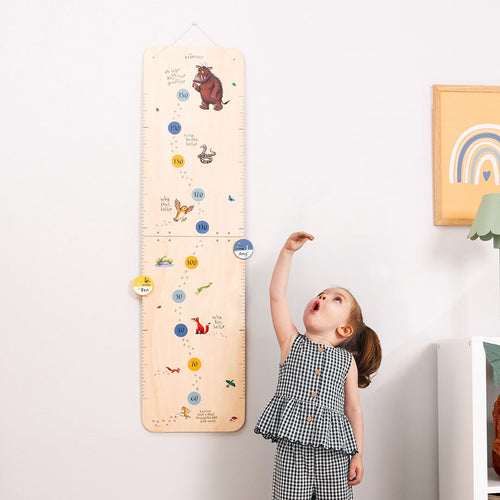 A young girl stands by the wall, reaching up to measure her height on the TP Toys The Gruffalo Wooden Height Chart - FSC® certified, featuring playful animal designs and numbers.