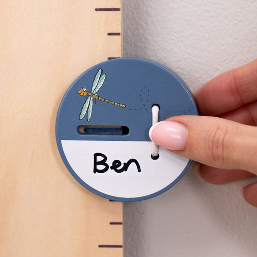 A hand adjusts a round, blue marker labeled Ben on The Gruffalo Wooden Height Chart - FSC® certified by TP Toys, decorated with a small dragonfly illustration.