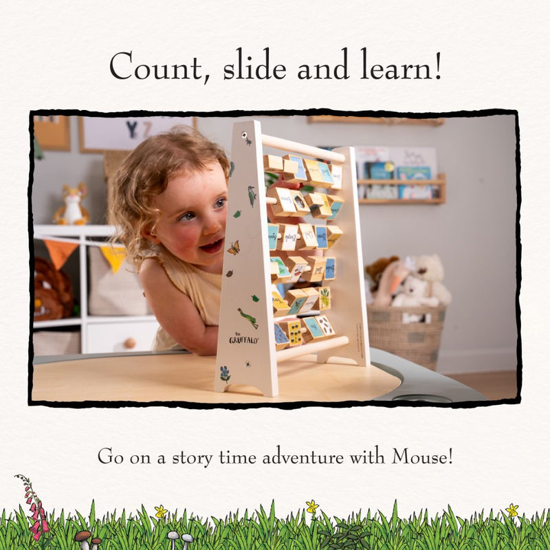 A young child smiles while playing with The Gruffalo Wooden Mini Abacus - FSC® certified by TP Toys at a table, an engaging early learning toy that helps develop fine motor skills. The background features a cozy room with stuffed animals and books.