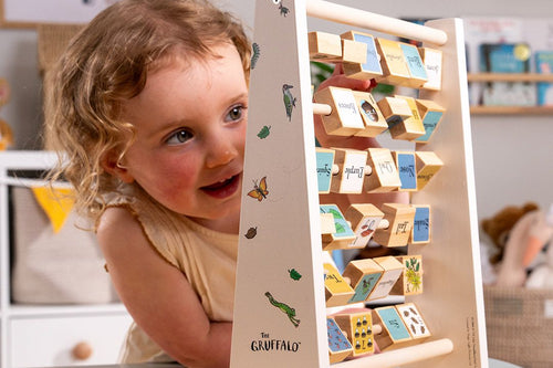 A young child plays with the TP Toys The Gruffalo Wooden Mini Abacus – an FSC® certified early learning toy designed to support fine motor skill development—surrounded by a playroom featuring illustrated alphabet blocks in the background.