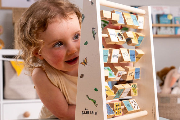 A young child plays with the TP Toys The Gruffalo Wooden Mini Abacus – an FSC® certified early learning toy designed to support fine motor skill development—surrounded by a playroom featuring illustrated alphabet blocks in the background.