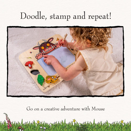A young child draws with a pen on The Gruffalo Wooden Magnetic Drawing Board - FSC® certified by TP Toys, surrounded by colorful illustrations on a light surface.