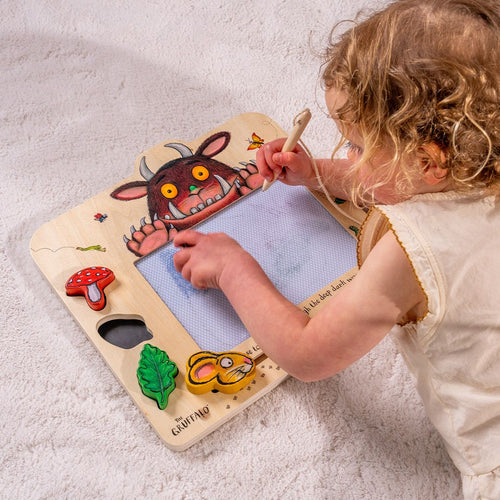 A young child draws with a pen on a The Gruffalo Wooden Magnetic Drawing Board - FSC® certified by TP Toys, seated on a light-colored carpet.